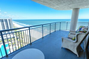Un balcón con vista al océano y la playa, una silla de mimbre y una mesa blanca.