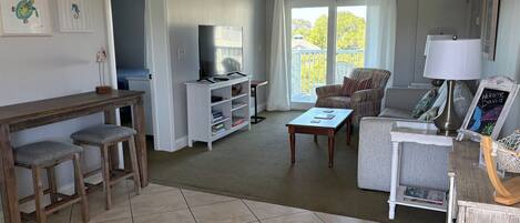 A living room with a TV, a coffee table, a sofa, and a chair. There is a balcony with a view of trees.