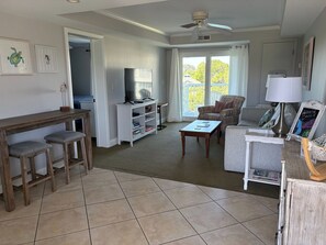 Un salon comprenant un téléviseur, une table basse, un canapé et un fauteuil. Il y a un balcon avec vue sur les arbres.