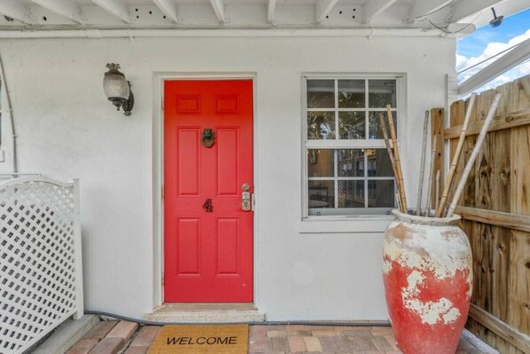 A red door with a number plate, a window with a view, and a welcome mat.