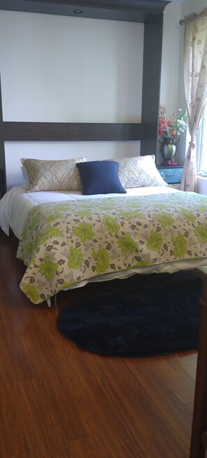 Un letto con una coperta a motivi floreali, un tappeto blu scuro e un comodino con un vaso di fiori.