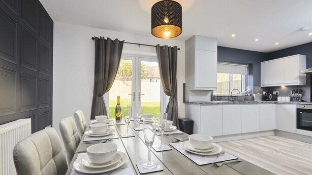 Un espace de repas moderne avec une longue table dressée pour six personnes, une cuisine aux meubles blancs et une vue sur une pelouse verdoyante, visible à travers de grandes fenêtres.
