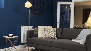 Un salon moderne avec un mur bleu foncé, un canapé gris, des coussins à motifs, une lampe sur pied et une petite table ronde sur laquelle repose un livre.