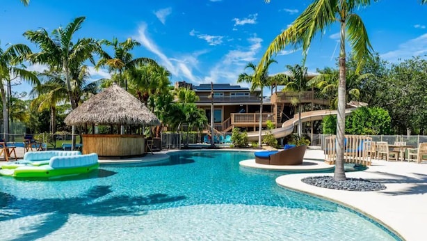 A resort pool area with a clear blue pool, lounge chairs, a thatched hut, and a palm tree. - 2 Home Sport Complex W/ Private Lake & Theater! (Jensen Beach)