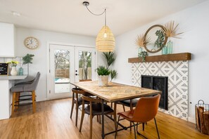 Uma sala de jantar com mesa de madeira, cadeiras, luminária pendente, relógio e lareira com ladrilhos estampados.