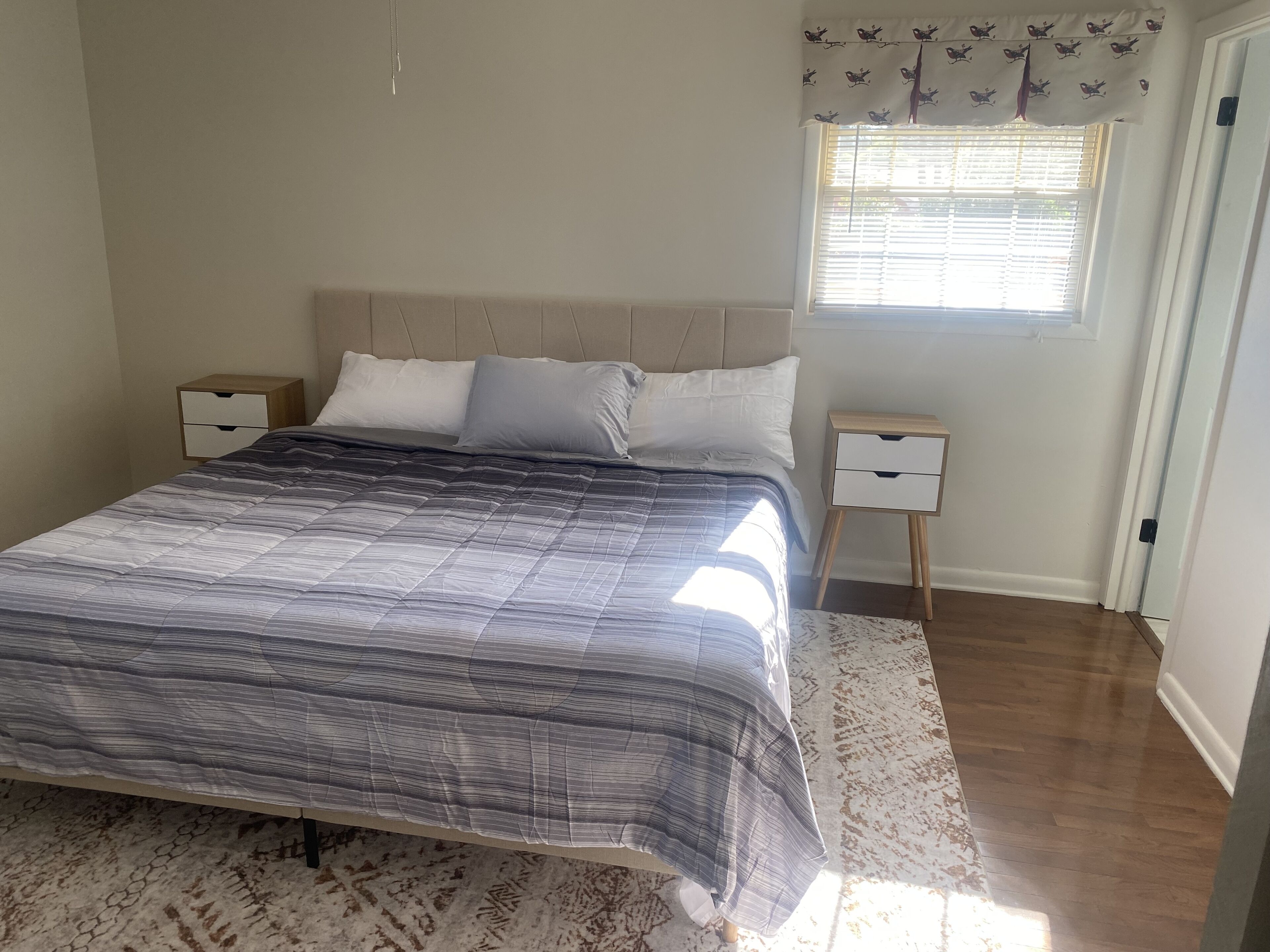 A bedroom with a bed, two bedside tables, a window with blinds, and a patterned rug.