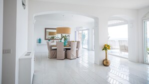 A spacious room with a white herringbone floor, a dining area with a table and chairs, and a large window offering a view of the sea. - Villa Biancalisa - Sea View Infinity Pool - Newly refurbished - Beachfront (Massa Lubrense)