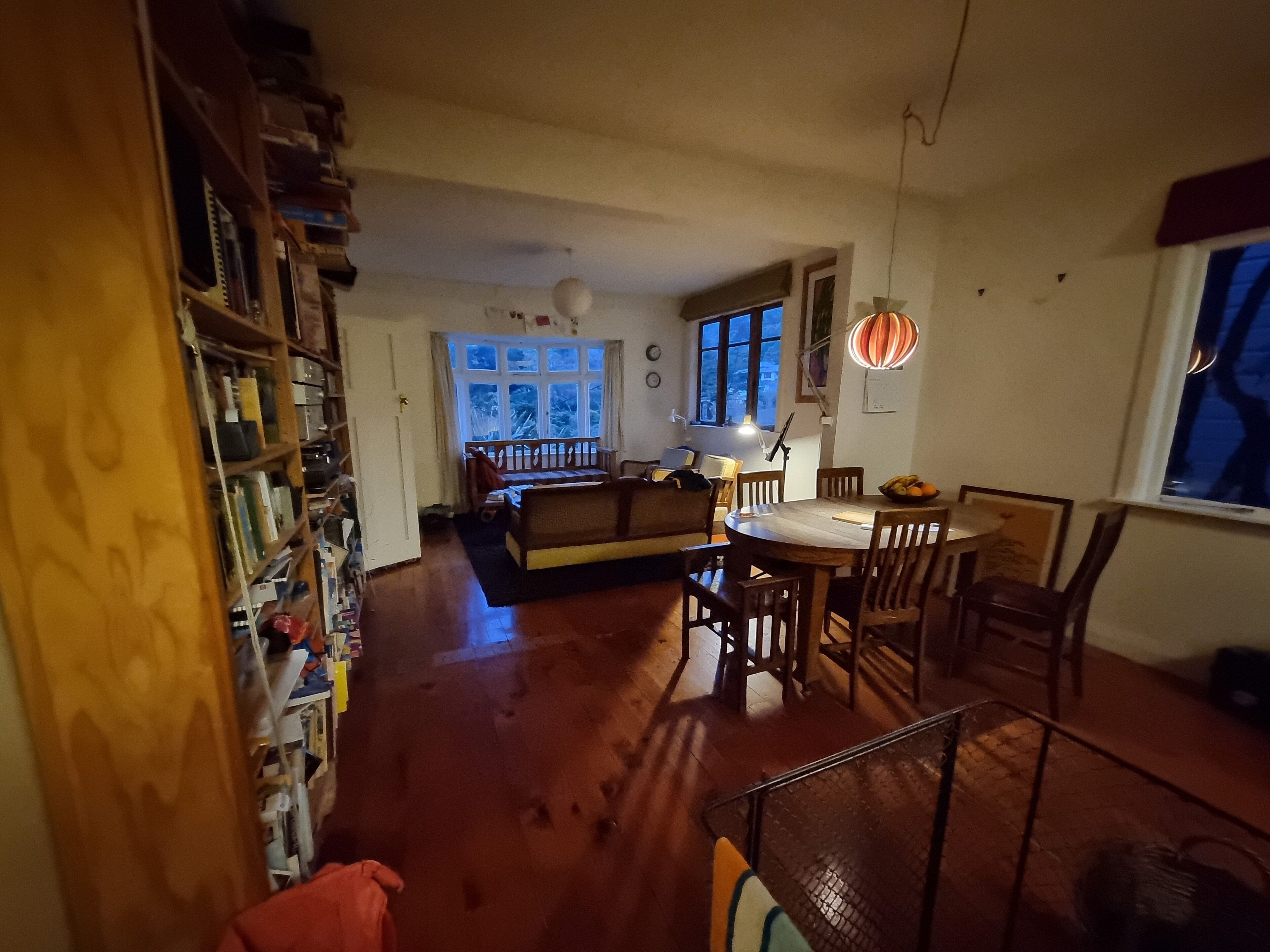 Un salon chaleureux avec un plancher en bois, une table à manger avec des chaises, un canapé et une bibliothèque remplie de livres et d’objets divers.