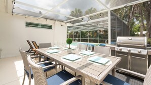 Un área de comedor al aire libre con cerramiento de vidrio, una parrilla y una piscina al fondo.