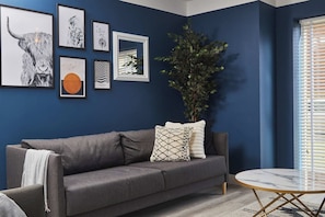 Uma sala de estar moderna com parede azul escura, sofá cinza, mesa de centro redonda e quadros enquadrados.