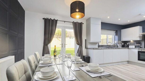 Een moderne keuken met witte kasten, een eethoek met een lange tafel voor zes personen, en uitzicht op een groen gazon door de ramen.