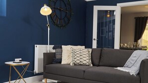 Een woonkamer met een donkerblauwe wand, een witte radiator, een moderne vloerlamp, een donkere bank en een klein, rond tafeltje met een lamp.