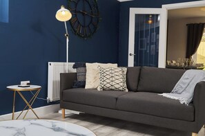 Sala de estar com parede azul escura, radiador branco, luminária de chão moderna, sofá escuro e mesa redonda pequena com uma luminária.
