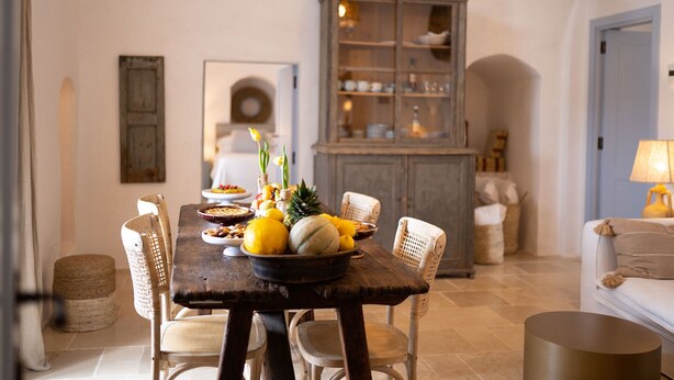 Comedor con mesa de madera dispuesta para una comida, rodeada de sillas de mimbre. En el fondo, un aparador con puertas de cristal y una lámpara a la derecha.