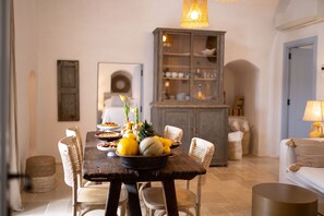 Comedor con mesa de madera dispuesta para una comida, rodeada de sillas de mimbre. En el fondo, un aparador con puertas de cristal y una lámpara a la derecha.