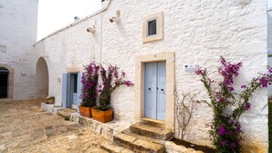 Un edificio blanco con puerta azul y una ventana pequeña, escalones de piedra y macetas con flores moradas.