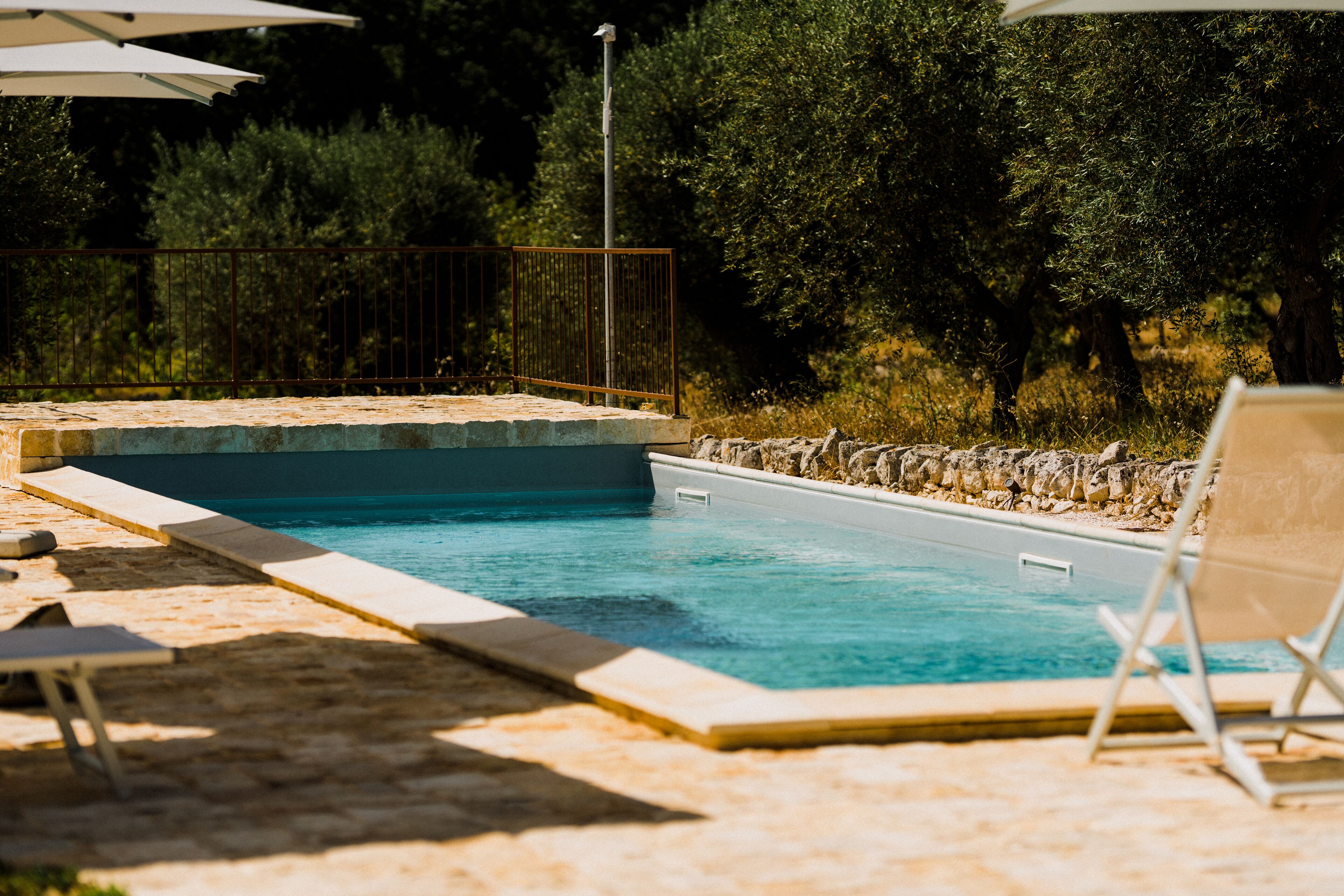 Una piscina con borde de piedra, rodeada por una zona de arena con sillas de descanso y sombrillas.