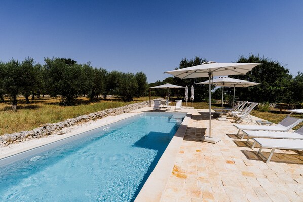 A swimming pool with lounge chairs and umbrellas, surrounded by trees and a stone wall. - Masseria Soluco (Monopoli)