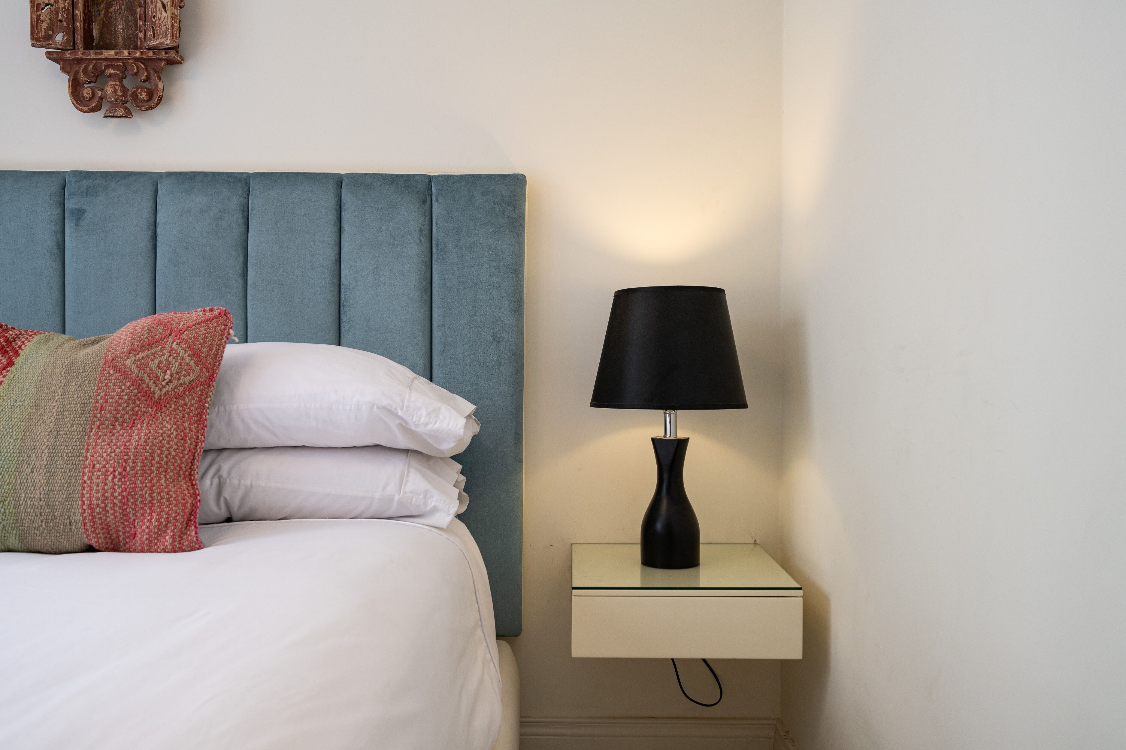 A neatly made bed with white linens and a red and green patterned pillow. A black lamp is on a small bedside table.