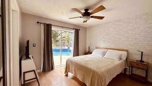 A bedroom with a bed, a ceiling fan, a TV on a stand, and a view of a swimming pool through a glass door. - Pirate’s Cove (Tarpon Springs)
