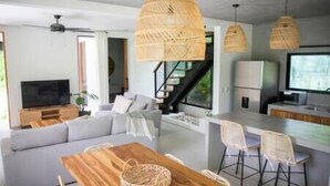 Ein modernes Wohnzimmer mit einer Couch, einem Holzesstisch und Korbstühlen. Eine Treppe führt zu einer oberen Ebene, es gibt eine Küchenzeile mit einem Kühlschrank und einem Fernseher.