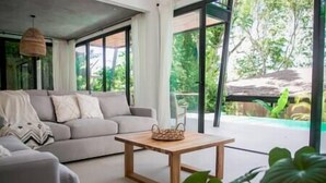 Ein modernes Wohnzimmer mit grauem Sofa, einem Holztisch und großen Fenstern mit Blick auf Grünflächen.