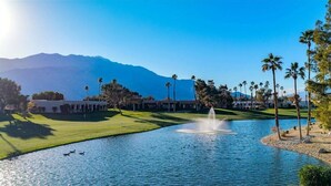 A golf course with a pond, palm trees, and mountains in the background.
