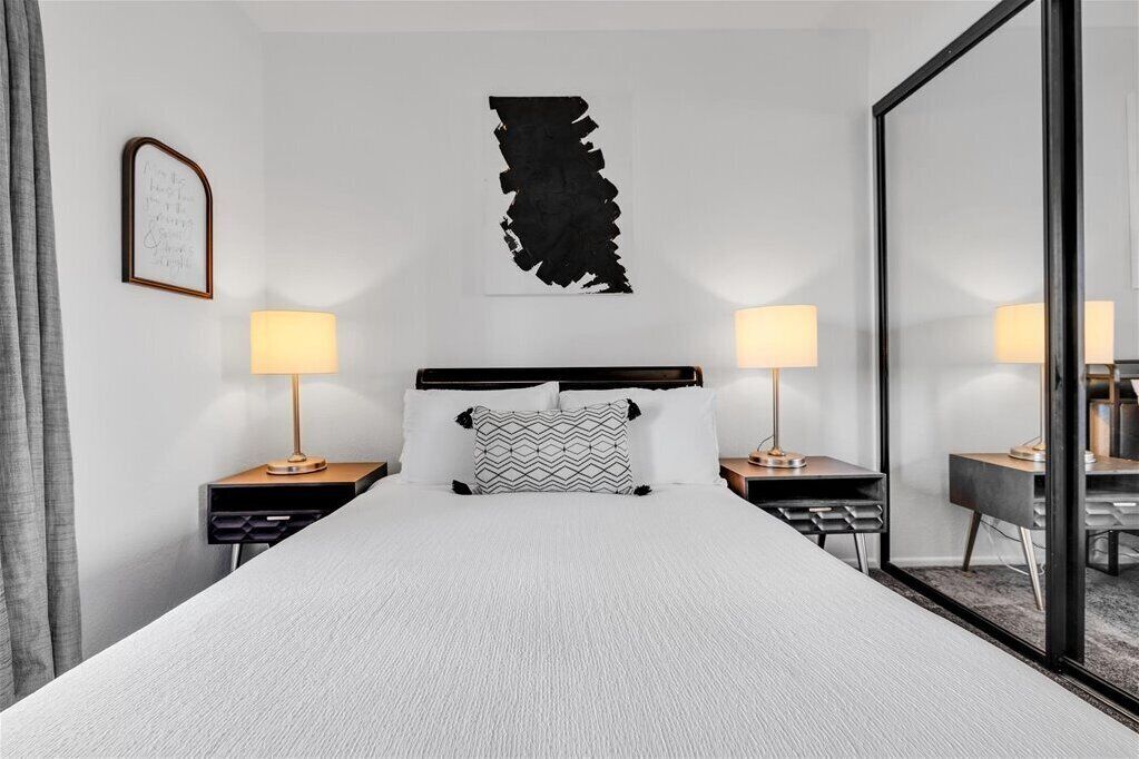 Et moderne hotelværelse med en stor seng, to sengeborde med lamper, et sort abstrakt kunstværk på væggen og et spejl til højre.