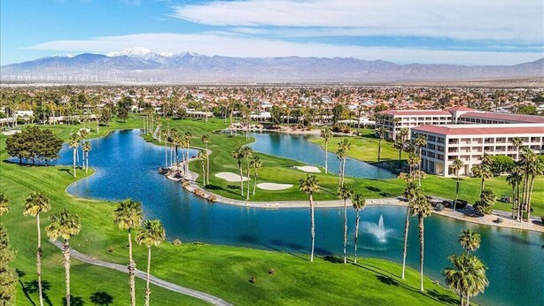 A golf course with multiple water features and palm trees, a large building with a red roof, and a mountainous backdrop.