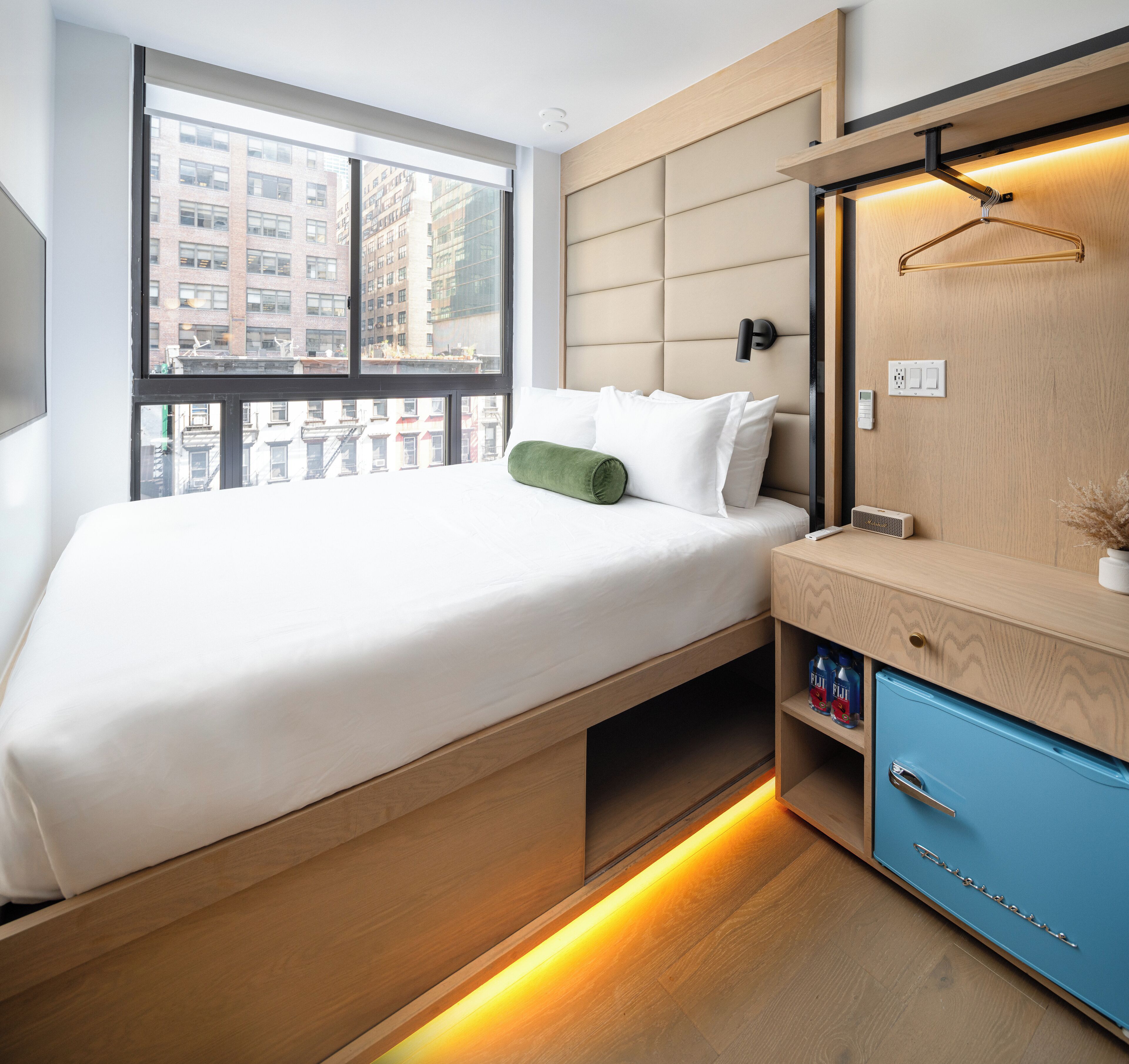 A modern hotel room with a bed, a wooden nightstand, a blue cabinet, and a large window offering a city view.