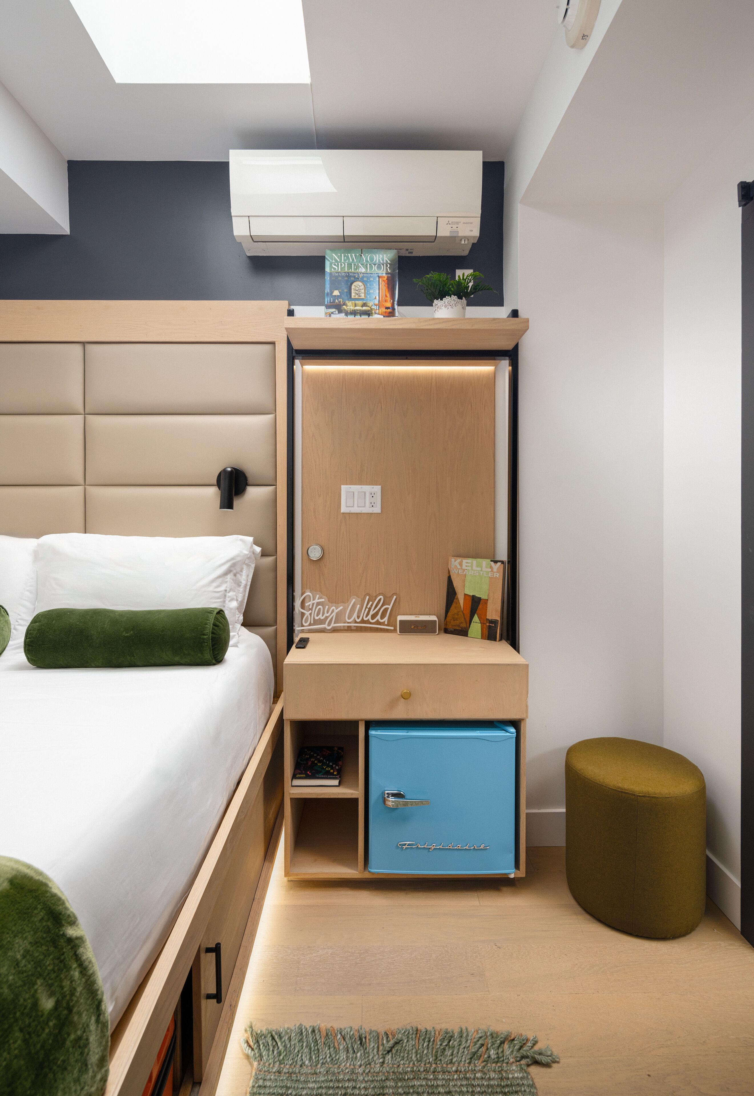 Quarto moderno com cama, mesa de cabeceira de madeira, móvel de armazenamento azul, almofada verde, um banquinho pequeno e um claraboia.