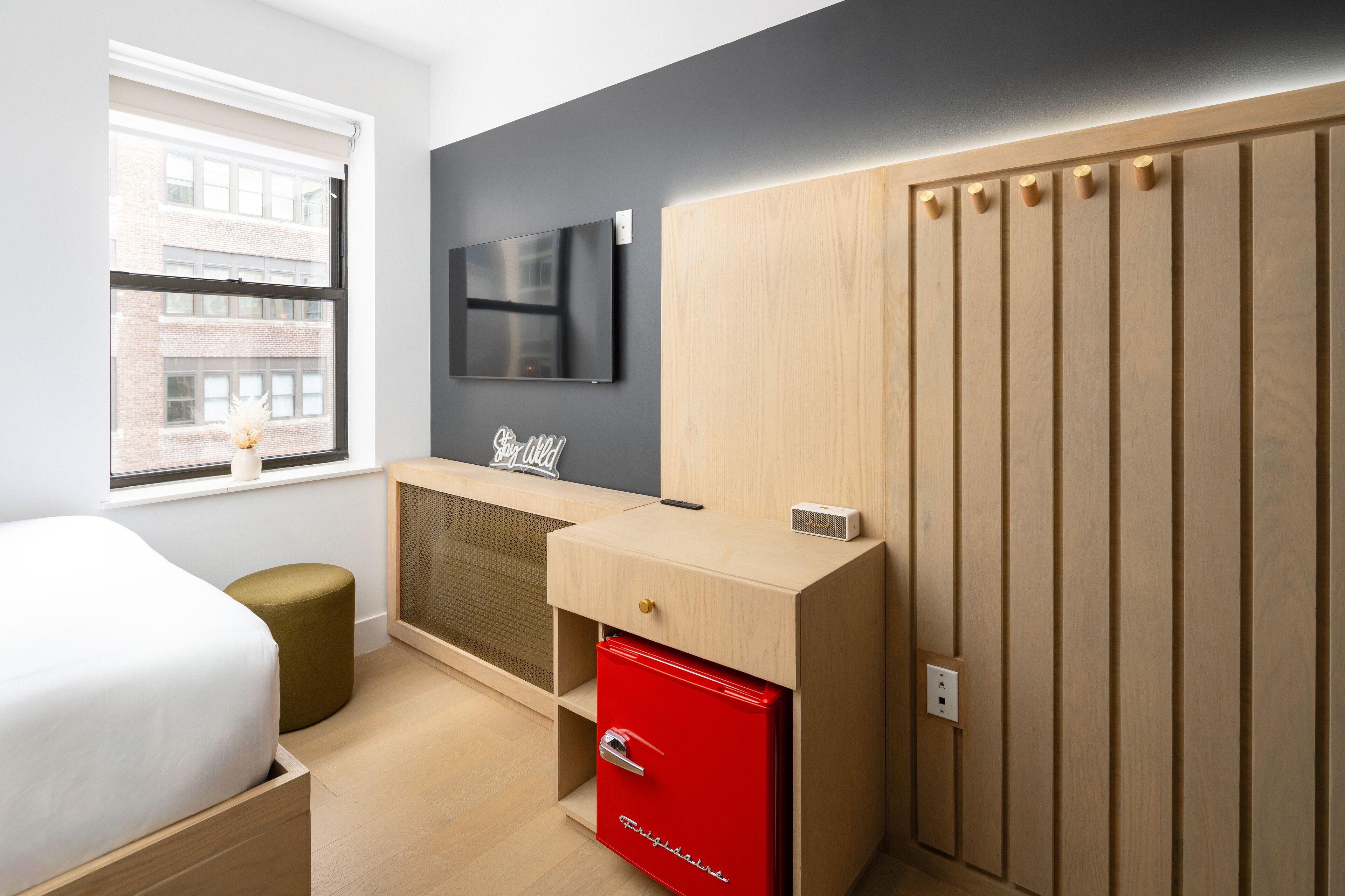 Un dormitorio moderno con una cama, un mueble de almacenamiento rojo, un televisor montado en la pared y una ventana con vistas a edificios.