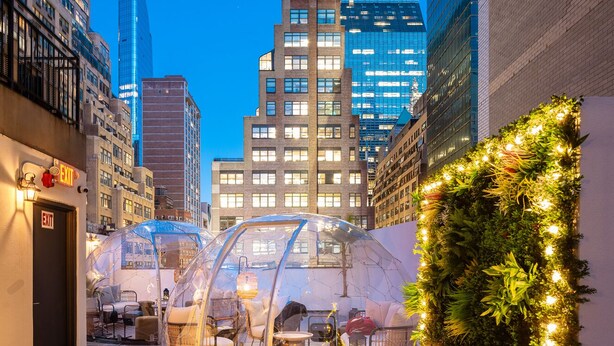 Una estructura de cúpula transparente con asientos en su interior, rodeada por un muro de follaje e iluminación, con el paisaje urbano al atardecer.