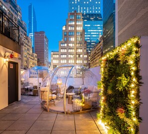 A transparent dome structure with seating inside, surrounded by a wall of greenery and lights, set against a cityscape at dusk. - Little Charlie Hotel (New York)