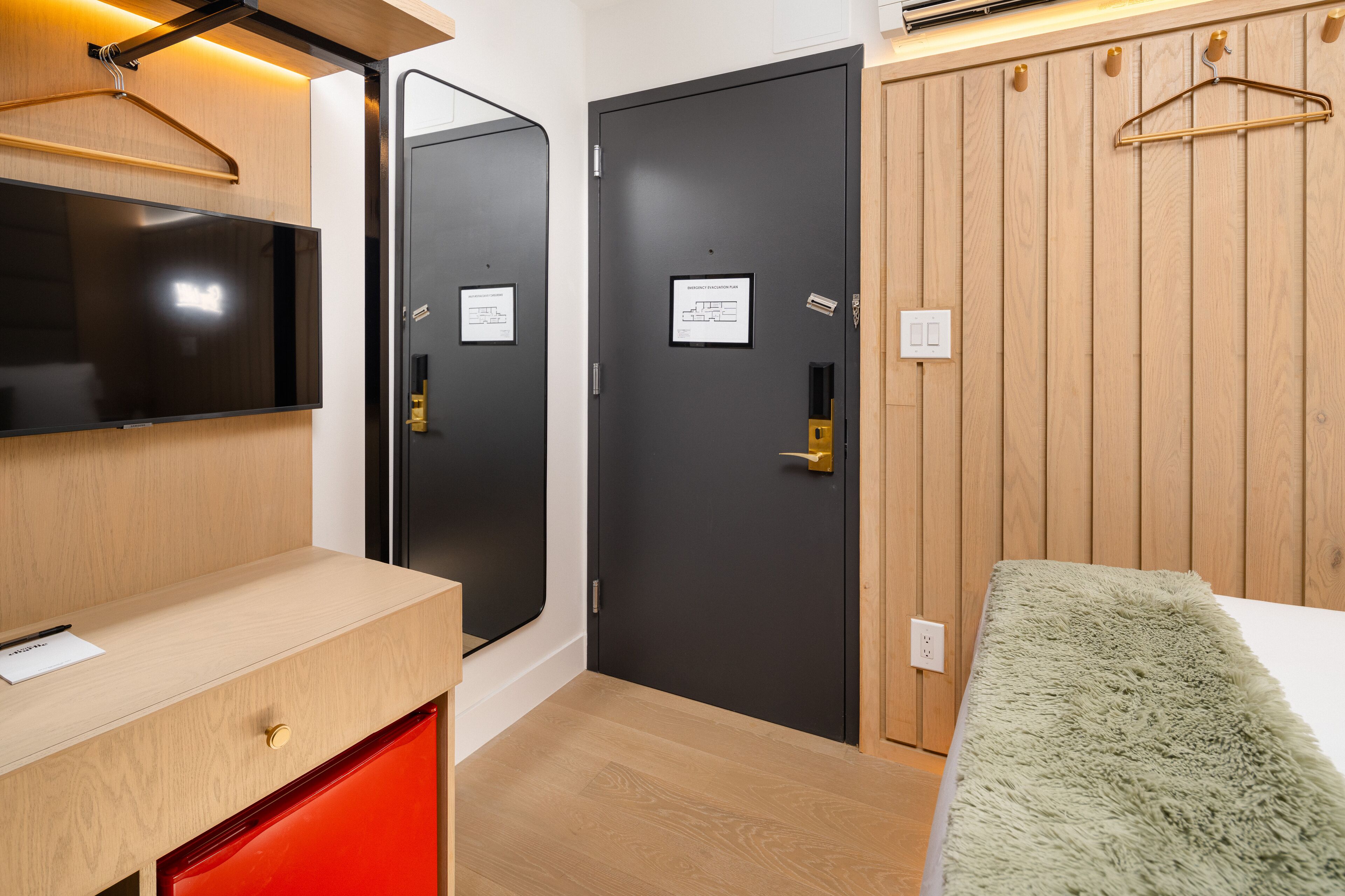 Una habitación moderna con un escritorio de madera, un televisor de pantalla plana, una puerta negra con cerradura de seguridad y una alfombra verde mullida.