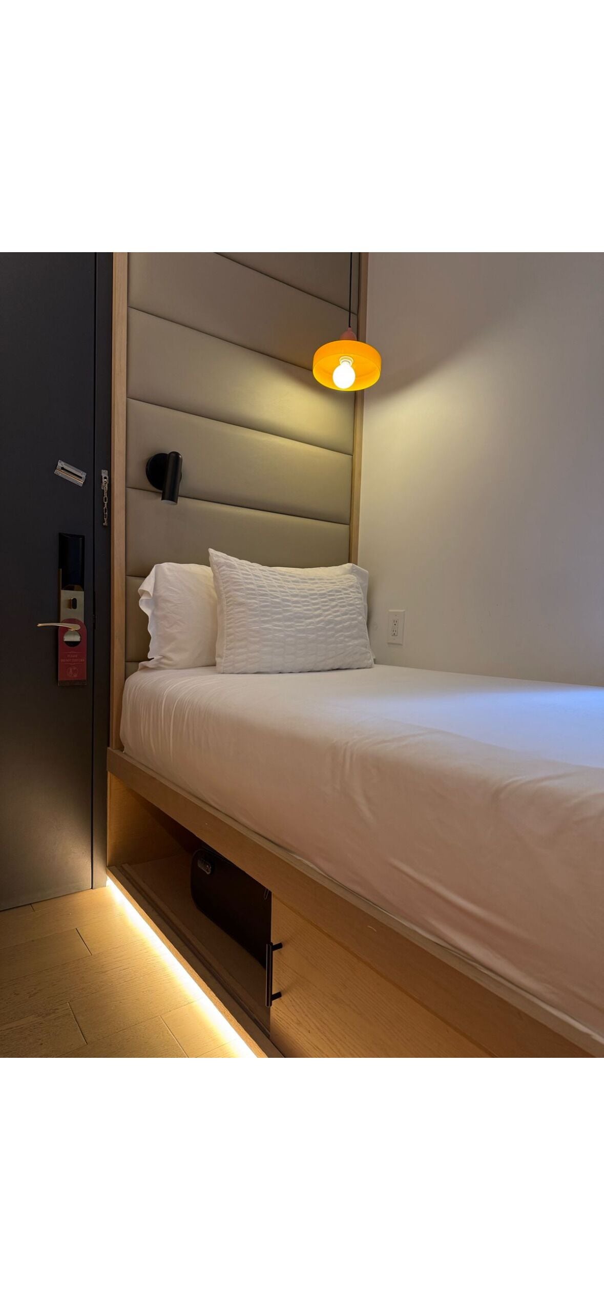 Uma cama de solteiro com cabeceira, uma luminária de cabeceira e uma gavetinha sob a cama.