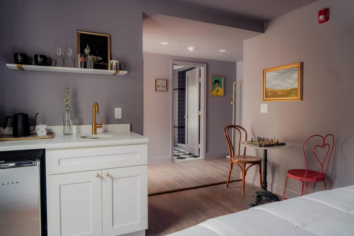 A modern hotel room with a white minibar, a wooden floor, a bed, a desk with a chessboard, and a red chair.