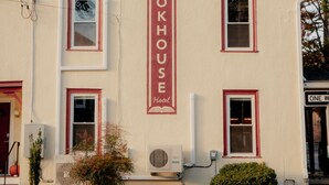 Un bâtiment à deux étages, doté d’une façade beige et de bordures rouges. On peut y voir une enseigne indiquant « BOOKHOUSE Hotel ». Il possède de nombreuses fenêtres, dont certaines sont équipées de volets, ainsi qu’un petit jardin avec des plantes et un mur de pierre à l’avant.