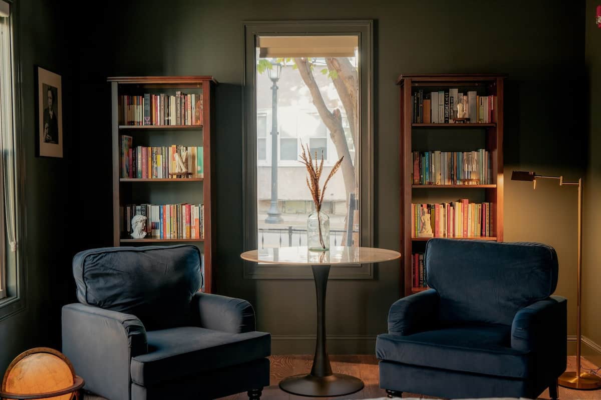 A room with two blue armchairs, a round table, and two wooden bookshelves filled with books.