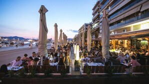 Un espace de restauration situé au bord de l’eau, avec des tables en plein air, des parasols et une vue sur le port de plaisance.