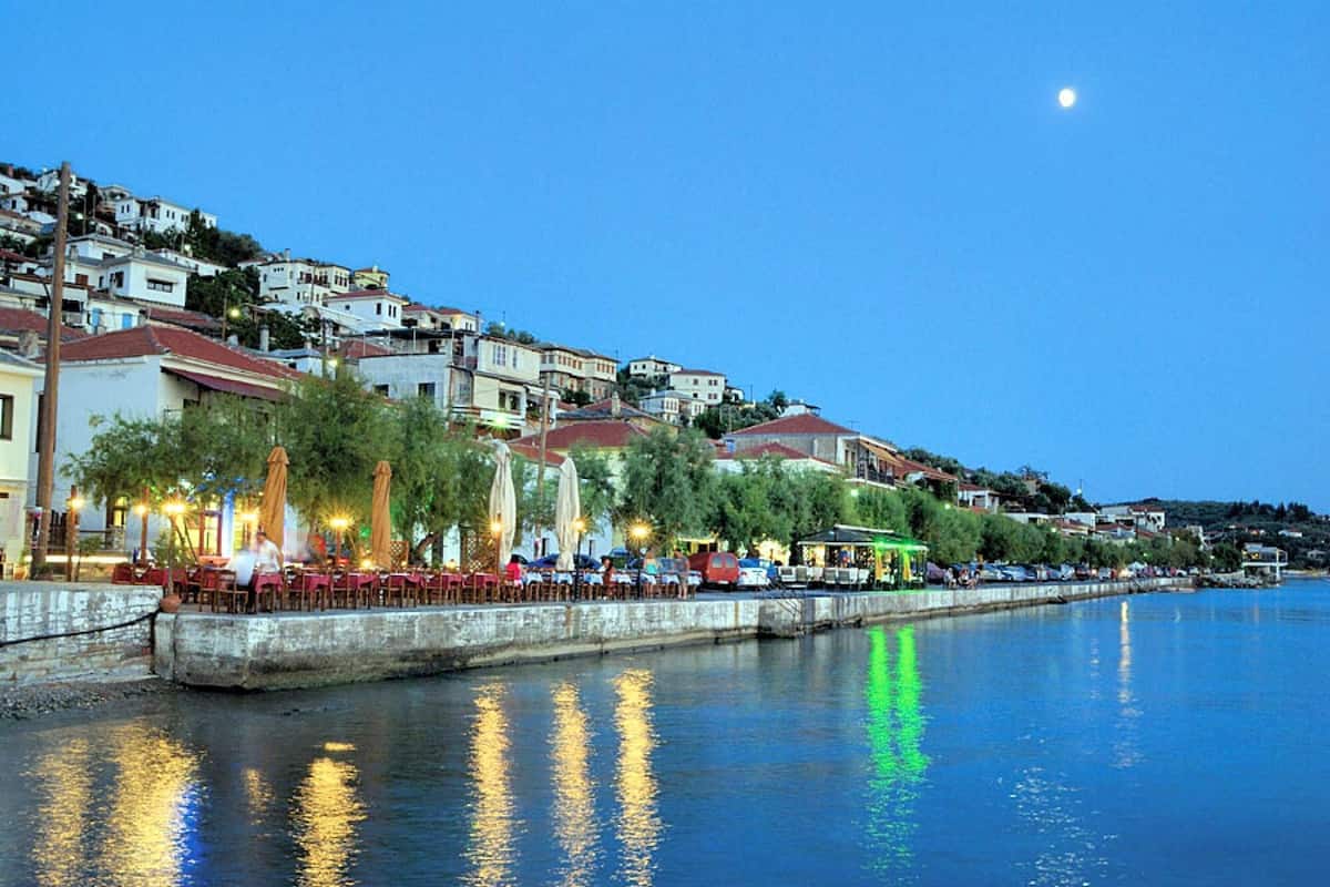 A waterfront promenade with buildings, trees, and a calm body of water reflecting lights.