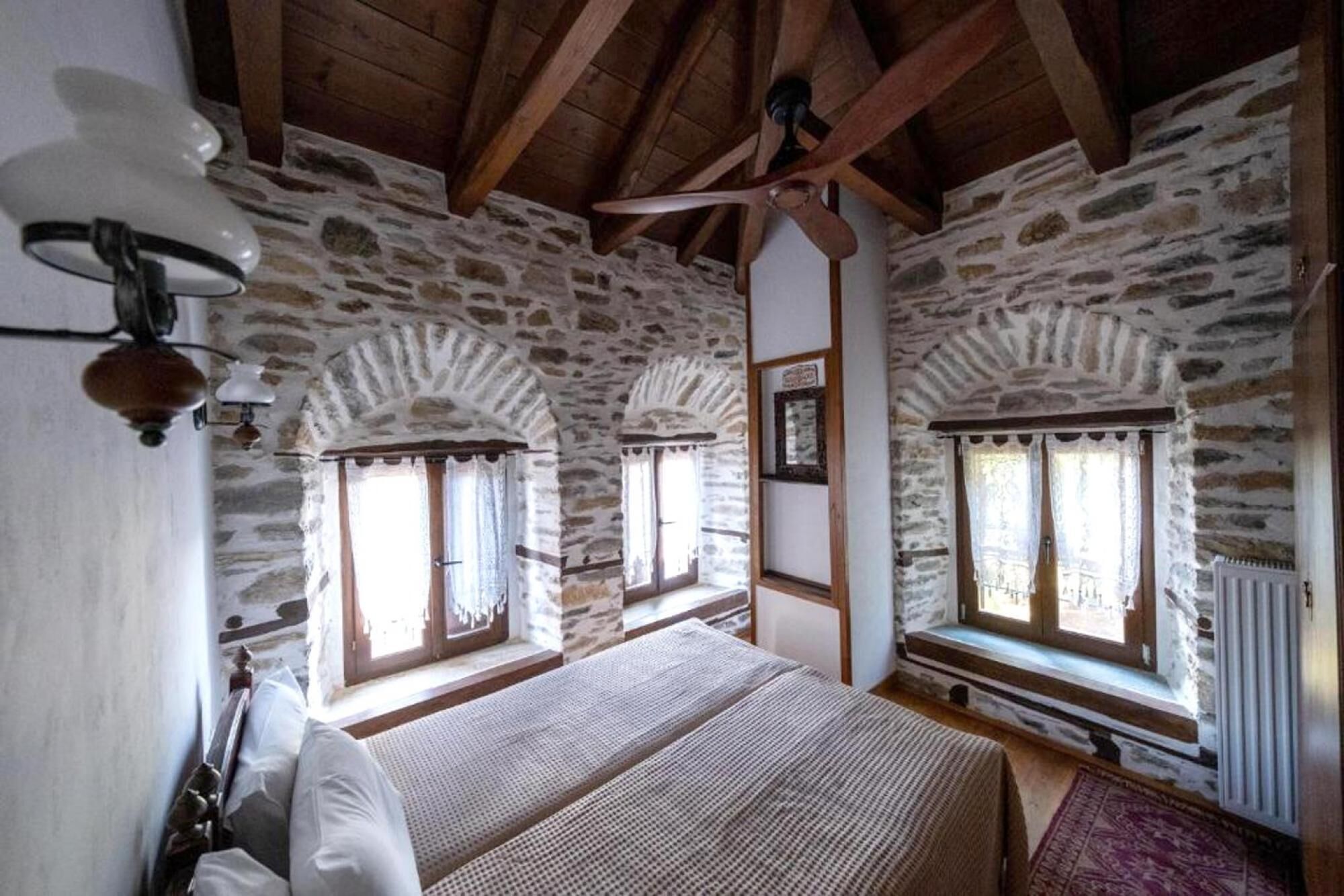 A bedroom with stone walls, wooden beams, and a ceiling fan. There are two windows with curtains and a bed with a blanket.