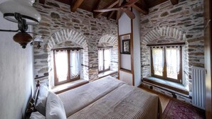 Une chambre avec des murs en pierre, des poutres en bois et un ventilateur de plafond. Il y a deux fenêtres avec des rideaux et un lit avec une couverture.