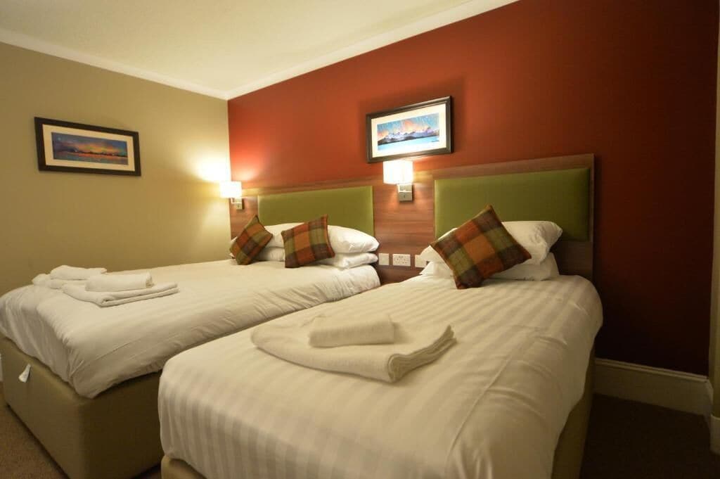 A hotel room with two beds, each with white linens and plaid pillows. There are wall-mounted lamps and a framed picture above the beds.
