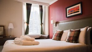 A hotel room with a bed, a nightstand, a lamp, a window with curtains, and a painting on the wall. - The Portree Hotel  (Portree)