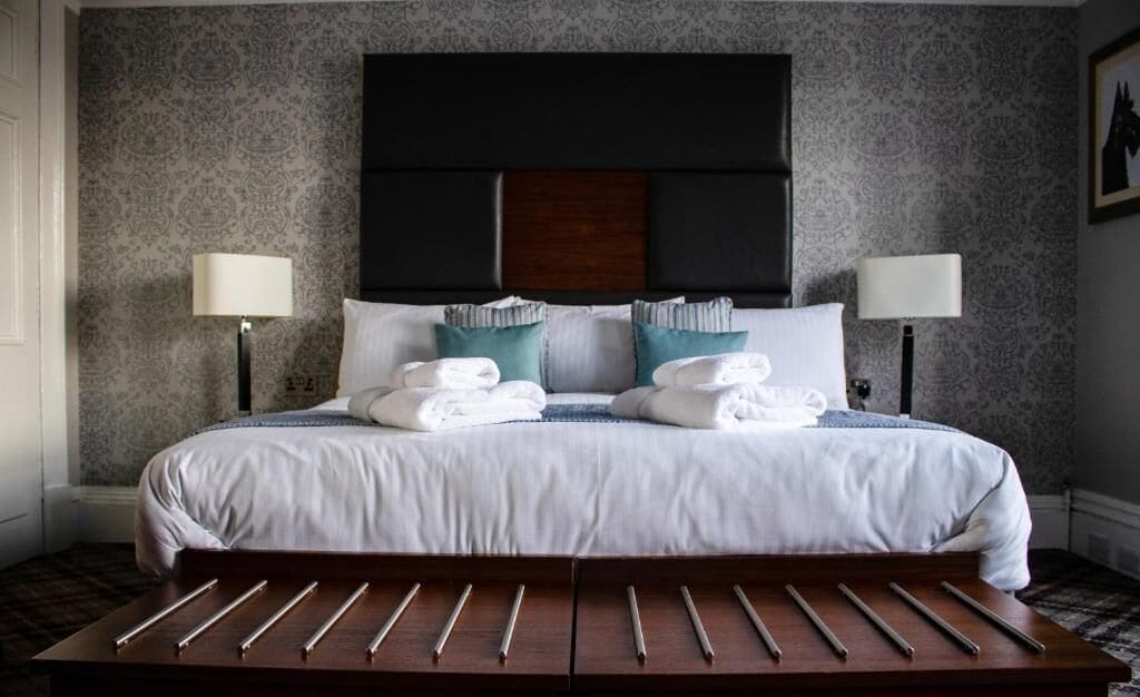 A neatly made bed with white linens and teal pillows, flanked by two bedside lamps.