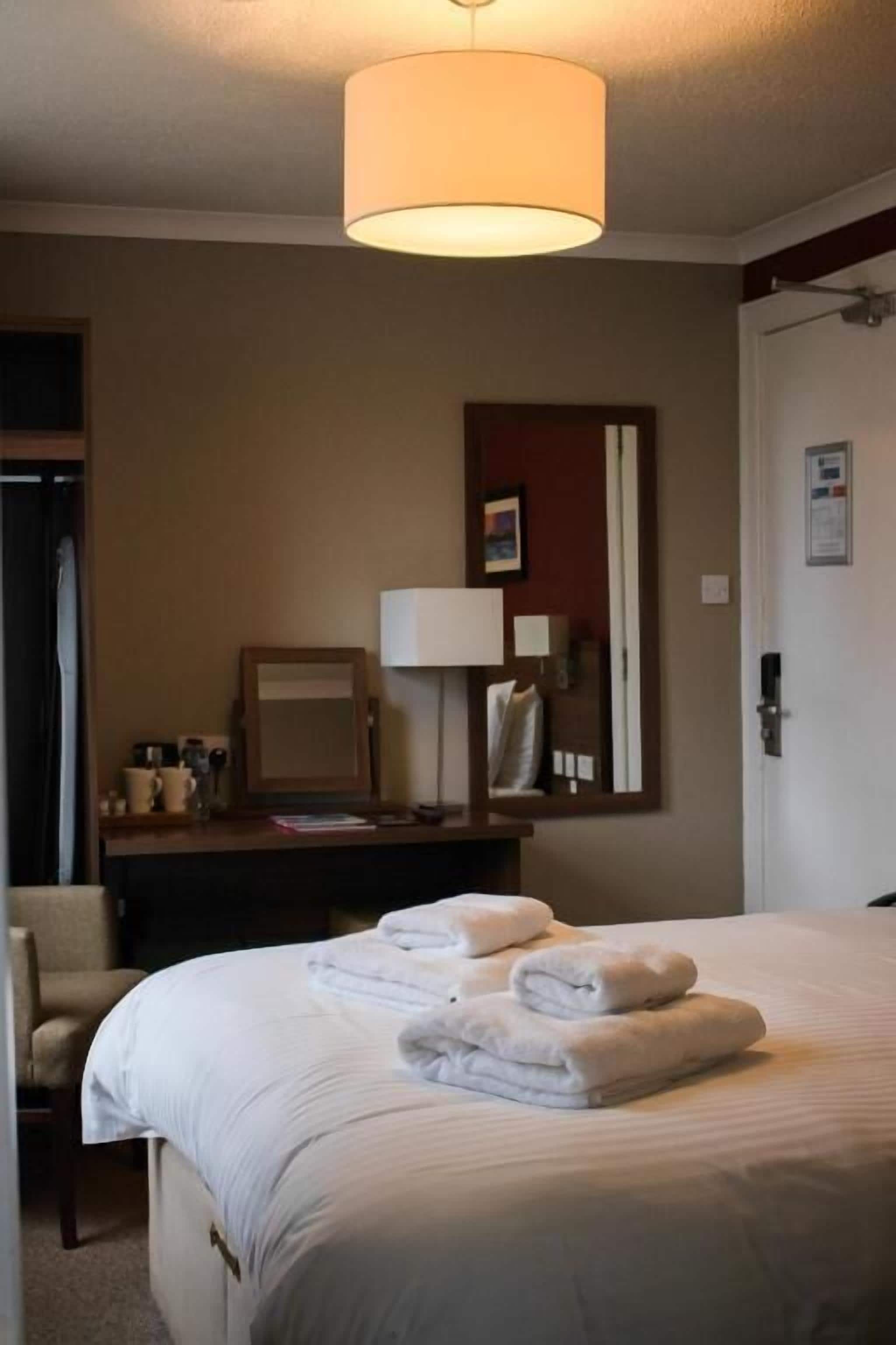 Quarto de hotel com cama, escrivaninha, cadeira, abajur e espelho.