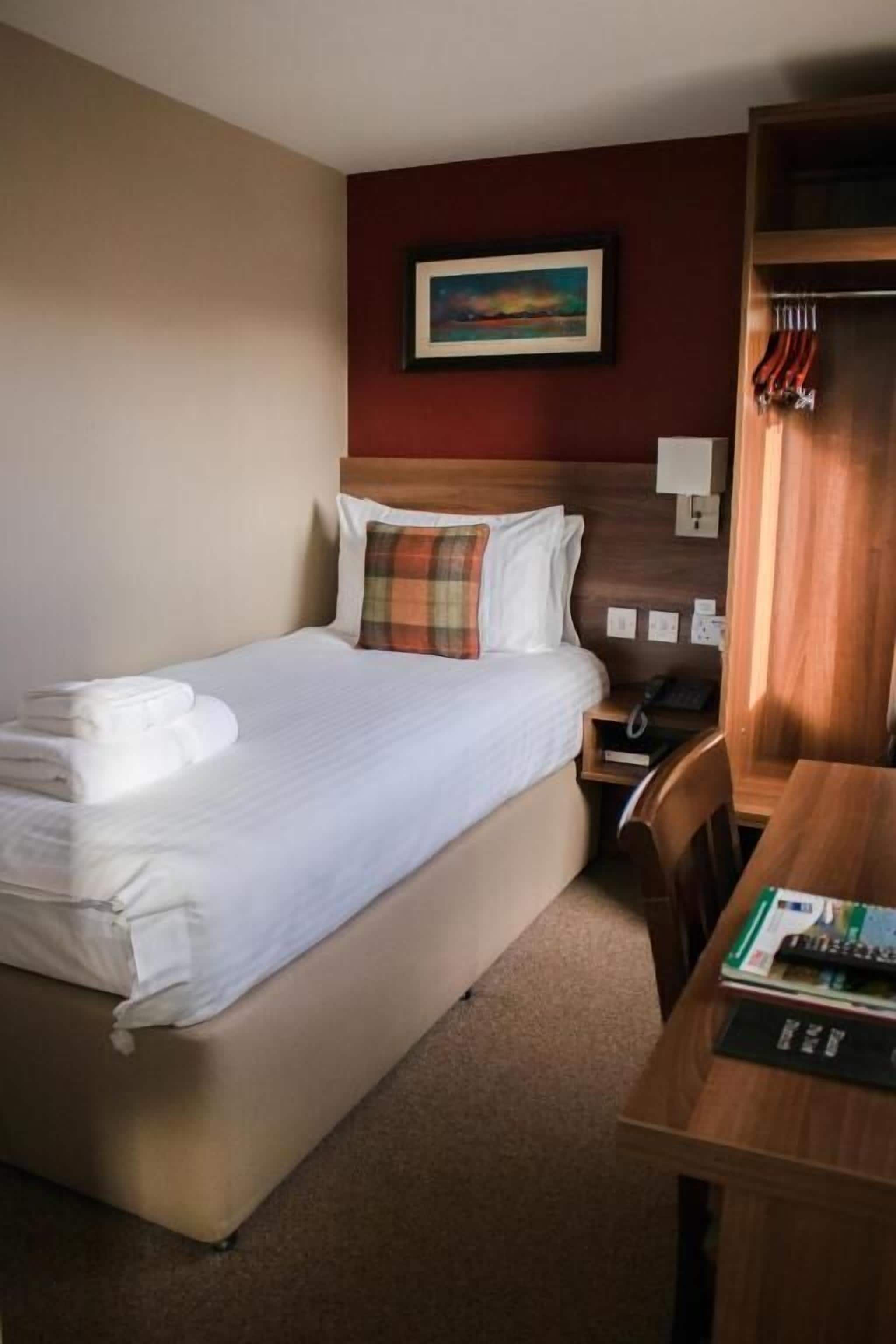 A hotel room with a bed, a desk, a chair, a wardrobe, and a painting on the wall.