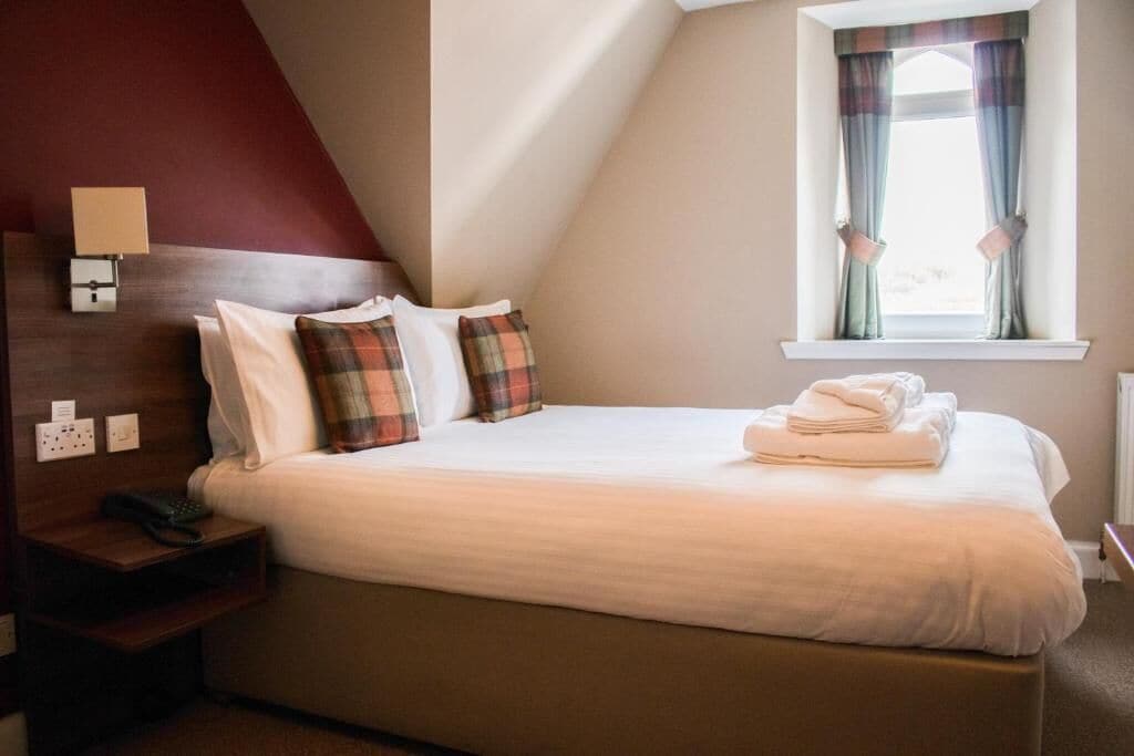 Une chambre d’hôtel avec un lit, une table de chevet, une lampe, une fenêtre avec des rideaux et un mur d’accent rouge.