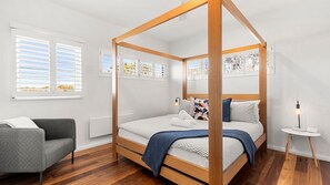 A four-poster bed with a wooden frame, a grey armchair, a bedside table with a lamp, and a view of the outdoors through the windows. - Ocean Dunes - Absolute Beachfront with Fireplace (Culburra Beach)